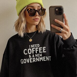 May include: Black sweatshirt with the text "I NEED COFFEE & A NEW GOVERNMENT" in white. The person in the image is wearing a neon green beanie, tortoiseshell sunglasses, and a gold necklace.