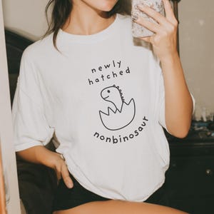 Könnte beinhalten: Weißes T-Shirt mit dem Text "newly hatched nonbinosaur" und einer Grafik eines Dinosauriers, der aus einem Ei schlüpft. Die Person auf dem Bild trägt eine rote Baseballkappe, eine schwarze Sonnenbrille und rote Cowboy-Stiefel.