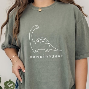 以下が含まれることがあります： 白い恐竜のイラストと「nonbinosaur」の文字が入ったオリーブグリーンのTシャツ。 恐竜は長い首と斑点のある体を持っています。 Tシャツは半袖です。