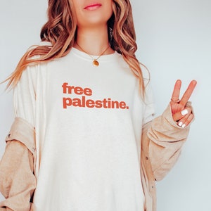 Puede incluir: Una camiseta blanca de gran tama&ntilde;o con el texto "free palestine" impreso en naranja en la parte delantera. La persona lleva una chaqueta de pana beige.