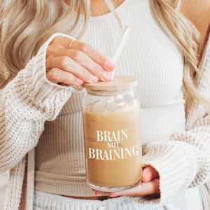 Pode incluir: Um copo de vidro transparente com tampa de madeira e canudo, cheio de uma bebida castanha clara. As palavras "BRAIN NOT BRAINING" estão impressas em branco na frente. O copo é segurado por uma pessoa que usa um suéter creme.