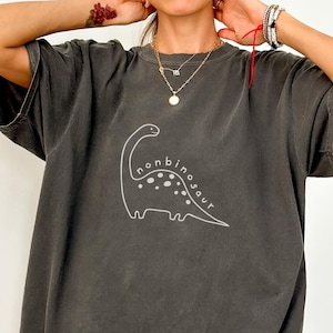 Może przedstawiać: Ciemnoszara koszulka z białym konturowym rysunkiem dinozaura i napisem "nonbinosaur". Koszulka ma krótkie rękawy i luźny krój. Grafika jest umieszczona na środku z przodu.
