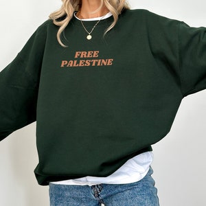 以下が含まれることがあります： 前面に茶色の「FREE PALESTINE」の文字がプリントされた濃い緑色のスウェットシャツです。