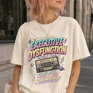 Puede incluir: Una camiseta de manga corta color hueso con un estampado gráfico que incluye el texto "Executive Dysfunction World Tour" y "Brain Fog Live". El diseño presenta un casete, planetas, estrellas y rayos en colores pastel.