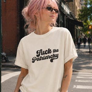 May include: An off-white short-sleeve t-shirt featuring the black text "Fuck the Patriarchy" in a bold, retro-inspired font. The shirt has a crew neckline and rolled sleeves, paired with light blue denim jeans.