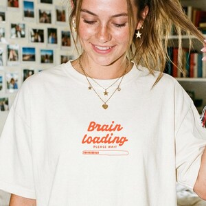 May include: A cream-coloured t-shirt featuring the text "Brain Loading" in red, with a loading bar graphic. The shirt is worn in a bedroom setting, paired with denim shorts. The room includes a bed, a bedside table, and a wall decorated with photographs.