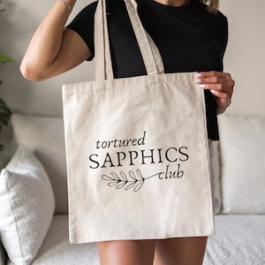 Puede incluir: Una bolsa de lona blanca con texto negro que dice "tortured SAPPHICS club" y un pequeño diseño floral negro.