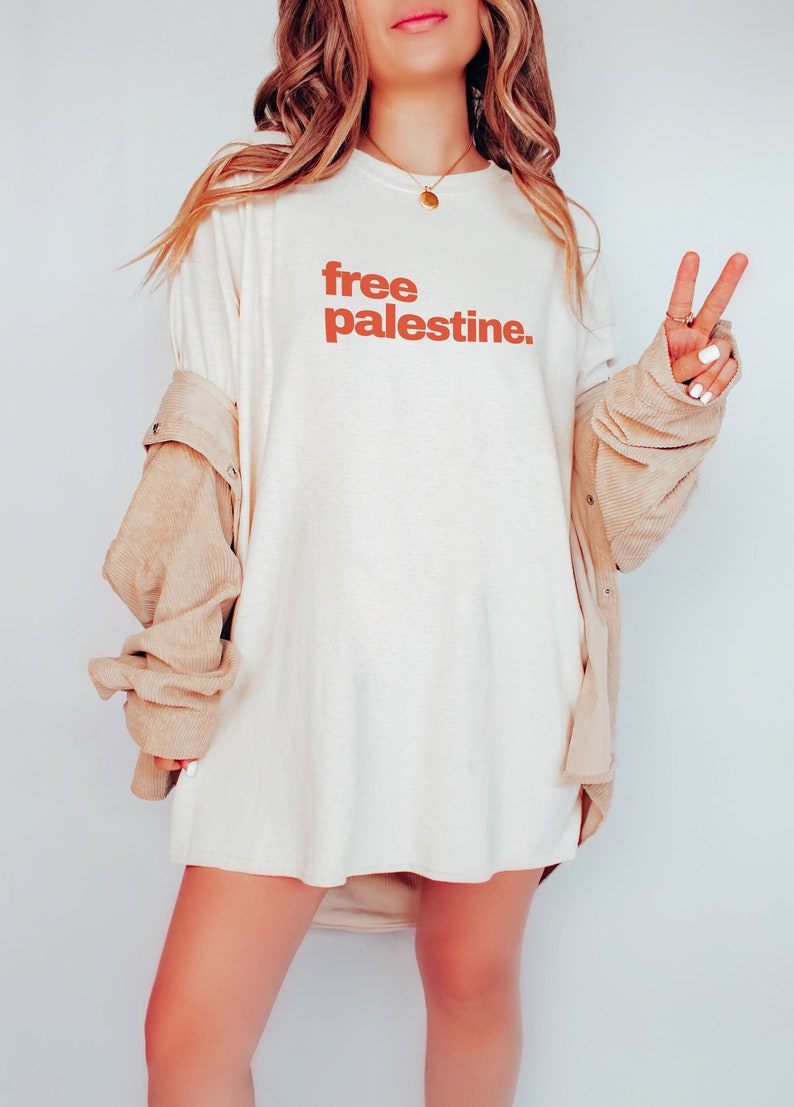 Puede incluir: Una camiseta blanca de gran tama&ntilde;o con el texto "free palestine" impreso en naranja en la parte delantera. La persona lleva una chaqueta de pana beige.