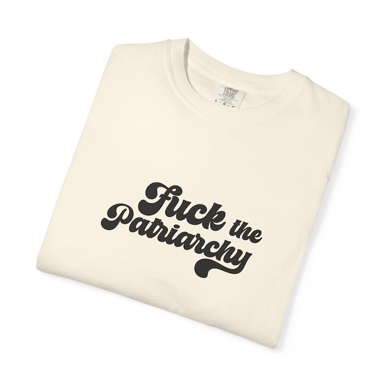 May include: A cream-colored t-shirt featuring the black text "Fuck the Patriarchy" printed in a bold, rounded, retro-style font across the chest. The garment has a crew neckline and a visible interior neck label indicating a size large.