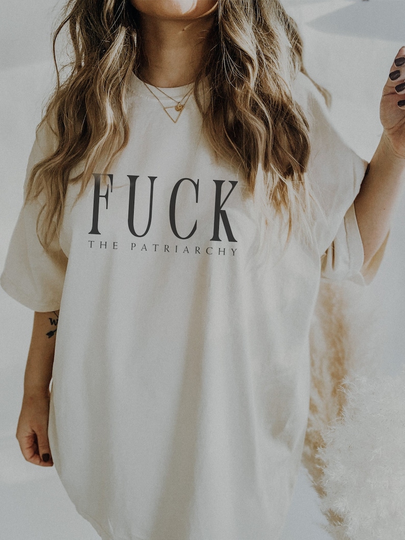 May include: Cream-colored t-shirt with the text "FUCK THE PATRIARCHY" in black, bold letters. The shirt has short sleeves and a relaxed fit. The image shows the upper body of a person wearing the shirt.