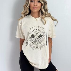 May include: A cream colored t-shirt with a black graphic print of a moth, mushrooms, and the word "sapphic".