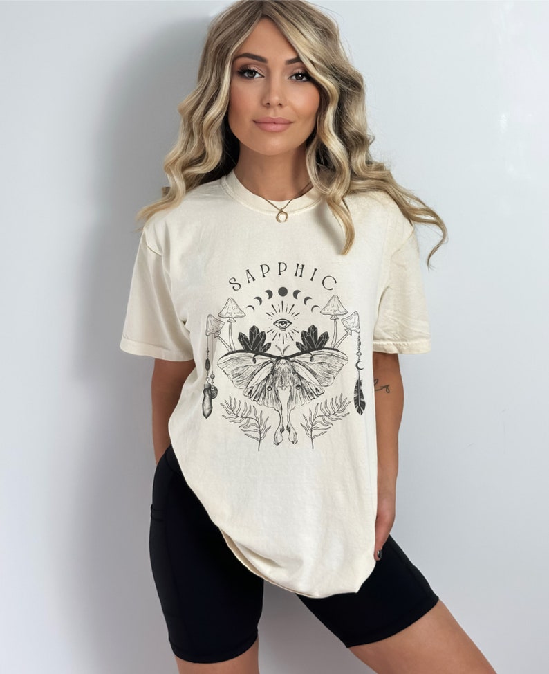 May include: A cream colored t-shirt with a black graphic print of a moth, mushrooms, and the word "sapphic".