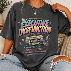 Op de afbeelding: Een antracietgrijs T-shirt met korte mouwen en een grafische print met de tekst "MY BRAIN PRESENTS EXECUTIVE DYSFUNCTION WORLD TOUR" en "BRAIN FOG LIVE". Het ontwerp bevat een cassettebandje, planeten en een lijst met zinnen.