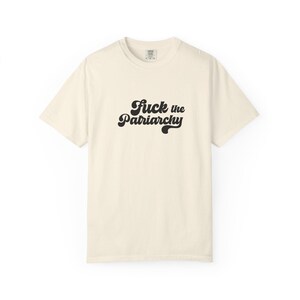 May include: A cream-colored short-sleeve t-shirt featuring the black text "Fuck the Patriarchy" printed in a bold, cursive, retro-inspired font across the center of the chest. The garment has a crew neckline and a standard, relaxed fit.