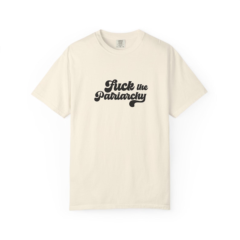 May include: A cream-colored short-sleeve t-shirt featuring the black text "Fuck the Patriarchy" printed in a bold, cursive, retro-inspired font across the center of the chest. The garment has a crew neckline and a standard, relaxed fit.