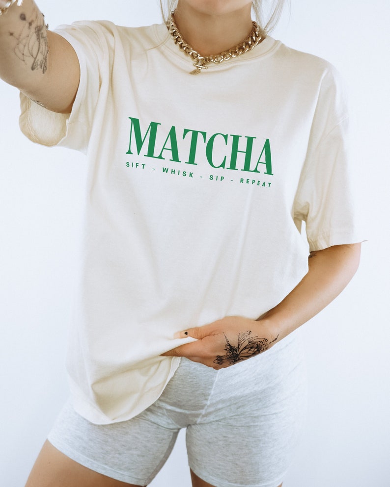 May include: Cream-colored t-shirt with the word "MATCHA" in bold green letters. Below, the shirt reads "SIFT - WHISK - SIP - REPEAT" in green. The shirt is a relaxed fit.