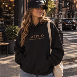 Pode incluir: Uma camisola preta com capuz apresentando o texto dourado SAPPHO e a citação someone will remember us no peito, combinada com um boné de basebol preto com texto branco new york e um saco de lona cor de creme sobre o ombro.