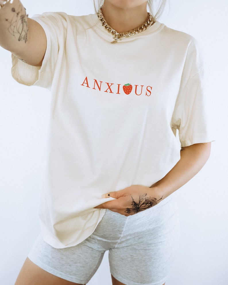 May include: A white oversized t-shirt with the word "ANXIOUS" printed in red with a red strawberry replacing the "I".