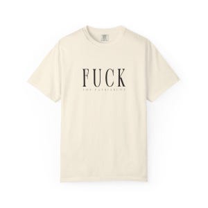 May include: Cream-colored t-shirt with the text "FUCK THE PATRIARCHY" in black, centered on the front. The text is in a bold, sans-serif font. The shirt has a crew neck and short sleeves.