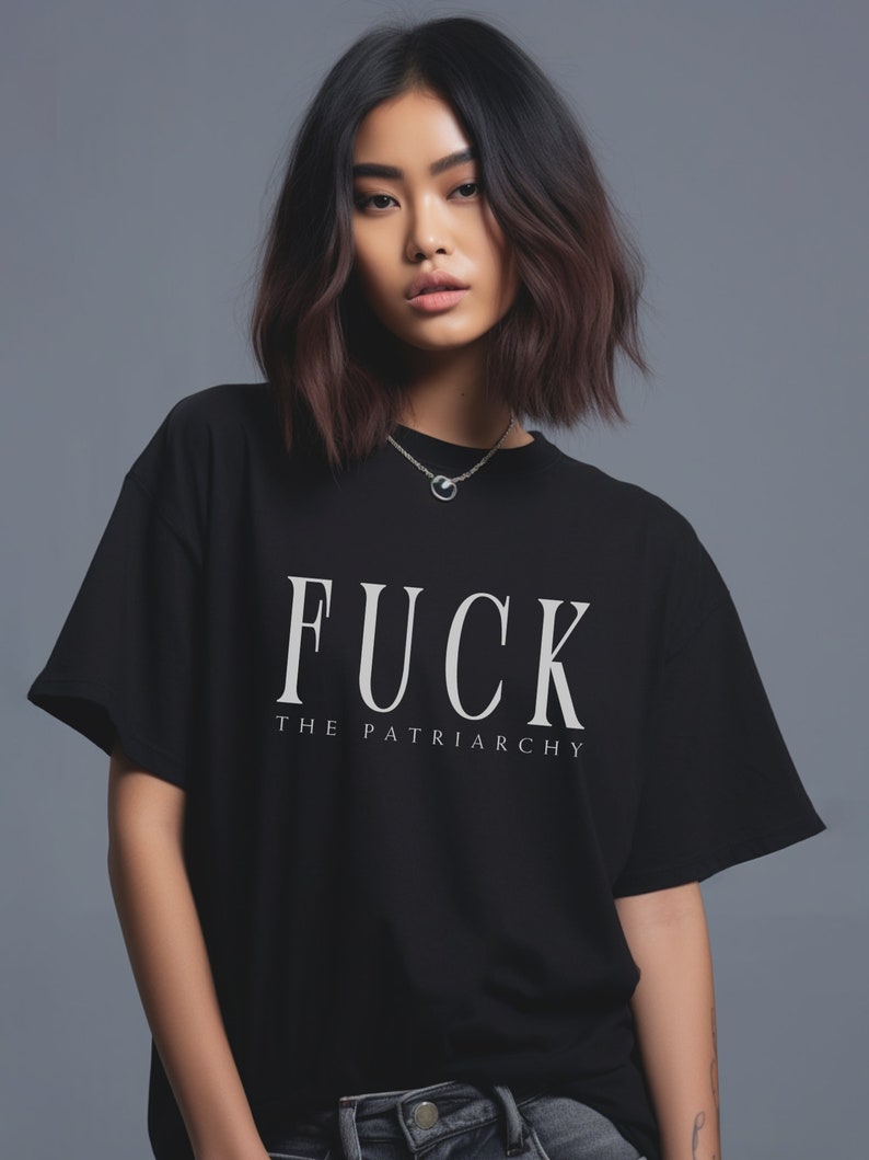 May include: Black t-shirt with the text "FUCK THE PATRIARCHY" in white, featuring a round neckline and short sleeves. The shirt is worn by a person with dark hair and a silver necklace.