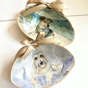 May include: Two decorative seashell dishes featuring hand-painted portraits of dogs. Each shell is adorned with a gold ribbon bow at the top and gold-painted edges. One shell depicts a dog on a beach, the other shows a dog named Nala.