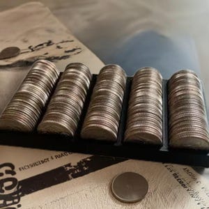 May include: A close-up of a black coin holder with five rows of silver coins. The holder is filled with stacks of coins, each row containing a different number of coins.