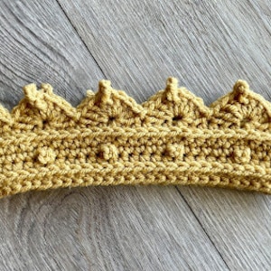 May include: A crocheted crown in a light yellow colour. The crown has a pointed top and a rounded base.