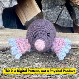May include: A crocheted mole toy in shades of purple, pink, and blue. The mole has a rounded body, small black eyes, and a pink nose. The wings are pink and blue. Text at the bottom reads: "This is a Digital Pattern, not a Physical Product."