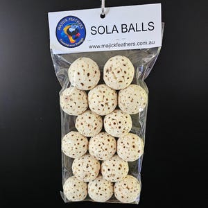May include: A transparent bag holds multiple off-white sola balls. Each ball features a textured surface with many small holes. The bag's header is white, displaying "SOLA BALLS" and a logo. The website address is also visible.