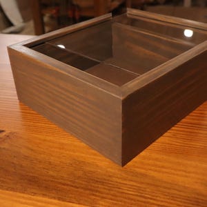 May include: A rectangular wooden box with a dark brown finish and a clear glass sliding lid. The box features a visible wood grain texture and a simple, clean-lined construction, providing a view into the interior compartment through the top.