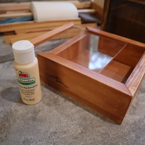 May include: A square wooden box with a hinged glass lid and a single internal divider, positioned next to a 2 fl oz bottle of cream-colored multi-surface satin acrylic craft paint labeled Toasted Marshmallow.