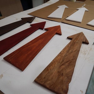 May include: A collection of flat, arrow-shaped wooden signs arranged in rows. The arrows feature various finishes, including dark brown, deep red, reddish-brown, natural wood grain, light blue, and solid white, all pointing in the same direction.