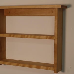 May include: A light brown wooden wall shelf featuring two horizontal tiers and a top ledge. The structure is composed of vertical side panels and horizontal shelves with visible screw holes on the upper back support bar for mounting purposes.