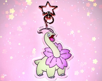 May include: A green and pink Pokemon keychain with a star-shaped charm. The Pokemon is a light green color with a pink flower around its neck. The keychain is made of clear acrylic.