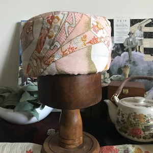 May include: A pink and gold fabric hat with a floral pattern, displayed on a wooden hat stand.