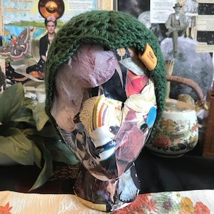 May include: A green crocheted beanie with a brown leather tag that says "Handmade". The beanie is on a mannequin head with a collage of images.