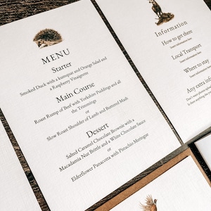 May include: A wedding menu card with a brown background and white text. The menu includes a starter, main course, and dessert. The starter is smoked duck with a kumquat and orange salad. The main course is roast rump of beef with Yorkshire puddings or slow roast shoulder of lamb and buttered mash. The dessert is salted caramel chocolate brownie with a macadamia nut brittle and a white chocolate sauce or elderflower panacotta with pistachio meringue.