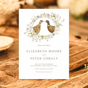 Pode incluir: Um cartão de convite de casamento branco com uma ilustração em aquarela de dois faisões dentro de uma coroa de flores. O texto diz "Save the date!" seguido de nomes e detalhes do casamento. O cartão está em uma superfície de madeira.