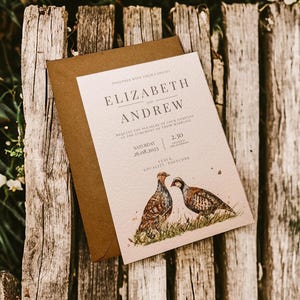 Puede incluir: Una invitación de boda con los nombres "Elizabeth y Andrew" impresos. La invitación presenta ilustraciones de acuarela de dos aves y un sobre marrón. La fecha es el sábado 26.08.2023. La hora es a las 14:30.