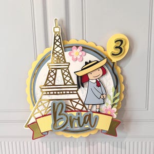 May include: A layered cake topper featuring the Eiffel Tower, a cartoon girl, and a balloon with the number 3. The name "Bria" is written on a banner. The design includes gold, yellow, blue, and pink accents.