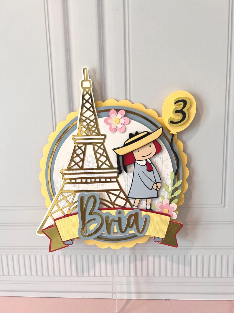 May include: A layered cake topper featuring the Eiffel Tower, a cartoon girl, and a balloon with the number 3. The name "Bria" is written on a banner. The design includes gold, yellow, blue, and pink accents.