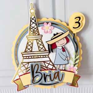 May include: A layered paper cake topper featuring a gold Eiffel Tower, a cartoon girl in a hat, and a yellow balloon with the number 3. The name "Bria" is displayed on a banner. Pink and yellow accents complete the design.