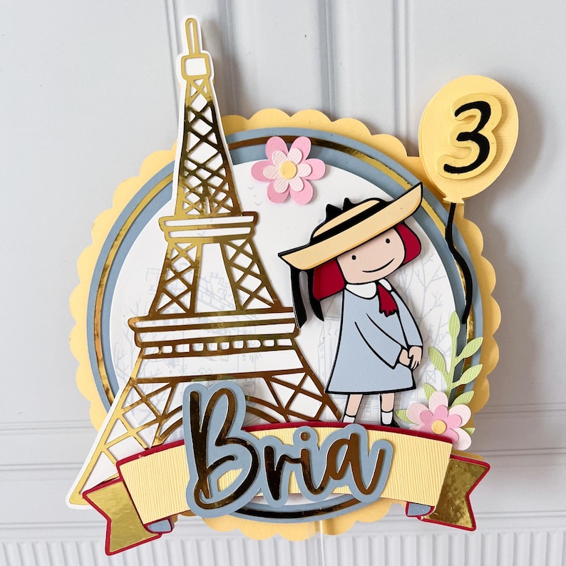 May include: A layered paper cake topper featuring a gold Eiffel Tower, a cartoon girl in a hat, and a yellow balloon with the number 3. The name "Bria" is displayed on a banner. Pink and yellow accents complete the design.