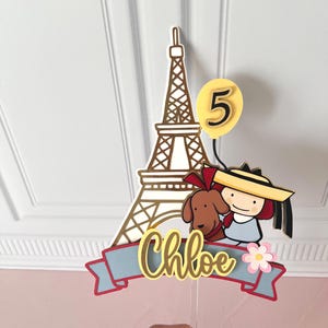 May include: A cake topper featuring a gold Eiffel Tower, a yellow balloon with the number 5, and a cartoon girl with a dog. The name "Chloe" is displayed on a banner with a pink flower.