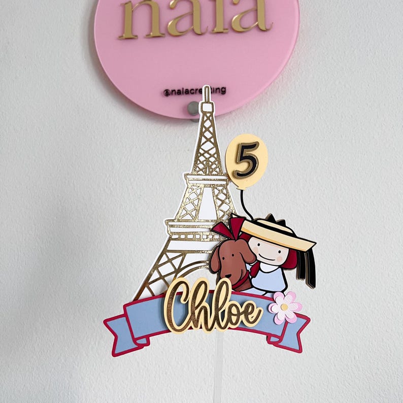 May include: A pink cake topper with gold lettering that says "nala" and "@nalacreting". The topper features a gold Eiffel Tower, a cartoon girl with a dog, and the name "Chloe" on a blue banner. A yellow balloon displays the number "5".
