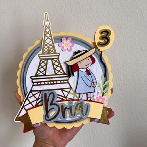 May include: A layered paper cake topper featuring a gold Eiffel Tower, a cartoon girl in a blue dress, and the name "Bria" on a banner. A yellow balloon with the number "3" is also included. Pink flowers and a blue and yellow circular design complete the festive look.