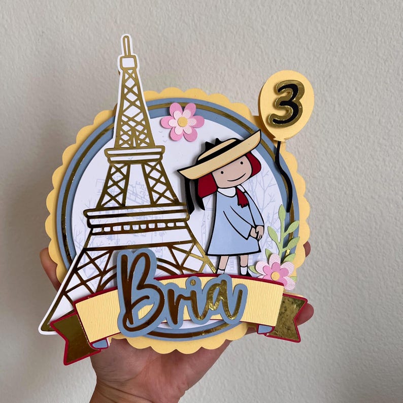 May include: A layered paper cake topper featuring a gold Eiffel Tower, a cartoon girl in a blue dress, and the name "Bria" on a banner. A yellow balloon with the number "3" is also included. Pink flowers and a blue and yellow circular design complete the festive look.