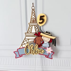 May include: A cake topper featuring the Eiffel Tower, a cartoon girl with a dog, and a number 5 balloon. The name "Chloe" is written on a blue banner with red trim. A pink flower is also included.