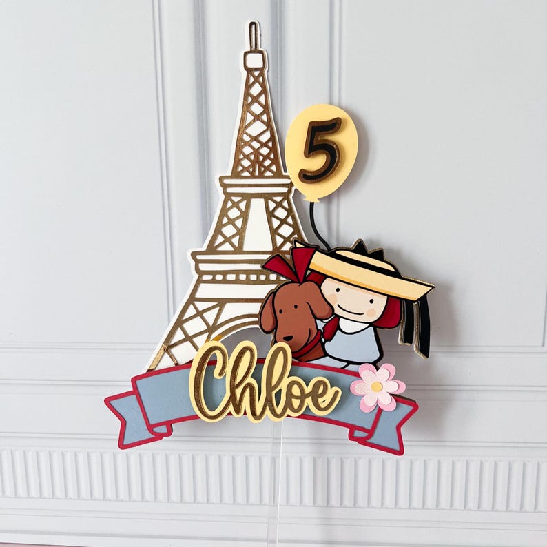 May include: A cake topper featuring the Eiffel Tower, a cartoon girl with a dog, and a number 5 balloon. The name "Chloe" is written on a blue banner with red trim. A pink flower is also included.