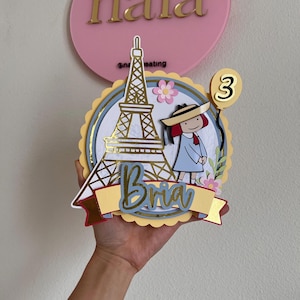 May include: A layered cake topper featuring a gold Eiffel Tower, a cartoon girl in a dress and hat, and a number 3 balloon. The topper includes the name "Bria" on a banner and is surrounded by flowers.